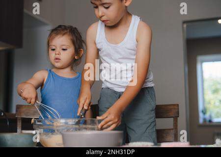Bruder und Schwester stehen am Küchentisch und mixen Zutaten für die Kuchentochter. Glückliche, fröhliche Familie. Konzept der Speisenrezepte. Stockfoto