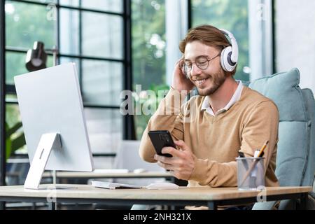 Ein lächelnder junger männlicher Programmierer, Freiberufler, sitzt im Büro am Tisch mit Kopfhörern und hält das Telefon in der Hand. Spricht bei einem Videoanruf, hört Hörbuch, Musik, Podcast. Stockfoto