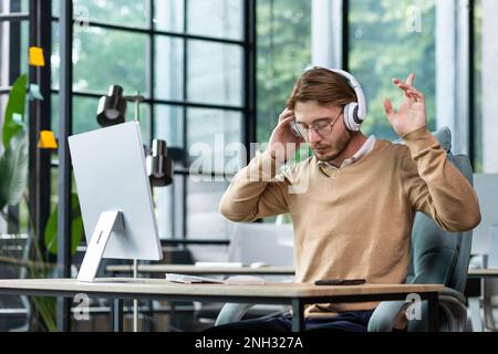 Ein junger männlicher Programmierer, Freiberufler, sitzt mit Kopfhörern im Büro und hört Musik. Er schloss die Augen, hob die Hände hoch, tanzte. Ruht sich aus, entspannt. Stockfoto