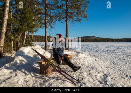 Eine ältere Frau ruht sich nach dem Skifahren aus, trinkt eine Tasse Kaffee und genießt die Sonne, Bild aus vasternorrland Schweden. Stockfoto