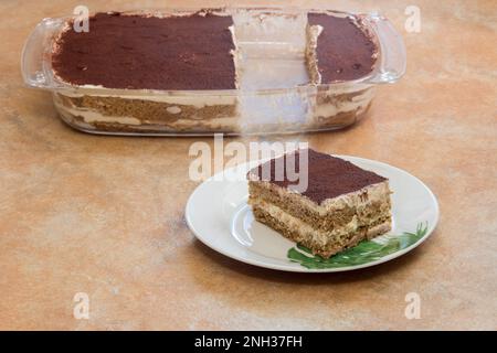 Schichten von cremigem Kakao, Kaffee und Käse in einem sichtbaren Glastablett mit hausgemachtem Tiramisu auf einer rustikalen Arbeitsfläche mit einem fehlenden frisch geschnittenen Stück Stockfoto