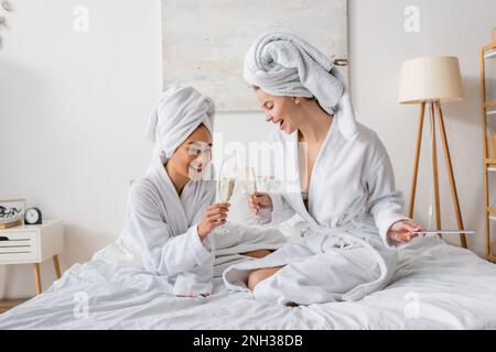 Fröhliche, gemischtrassige Frauen in weißen, weichen Bademänteln und Handtüchern, die Champagnergläser auf dem Bett klimpern, Stockbild Stockfoto