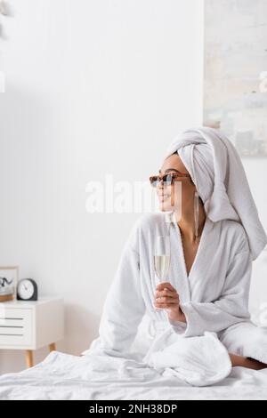 Lächelnde afroamerikanische Frau in Frotteebademänteln und trendigen Sonnenbrillen, die Champagner hielt und im Schlafzimmer wegschaute, Stockbild Stockfoto