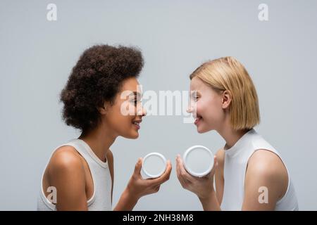 Seitenansicht von multiethnischen blonden und brünetten Frauen, die Kosmetikcreme halten und einander anlächeln, isoliert auf grauem Stockbild Stockfoto