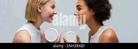 Seitenansicht von blonden und brünetten Frauen mit aneinander lächelnden Körpercreme, isoliert auf grauem Banner, Stockbild Stockfoto