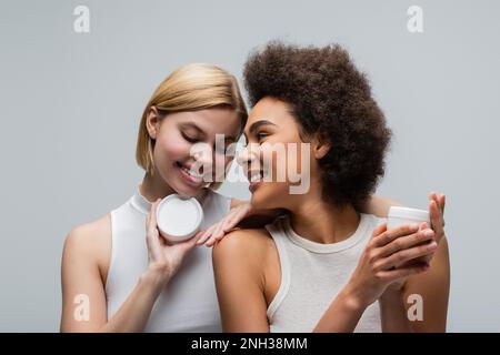 Junge und hübsche, multiethnische Frauen in weißen Tops, die mit Gläsern mit Körpercreme posieren, isoliert auf grauem Stockbild Stockfoto