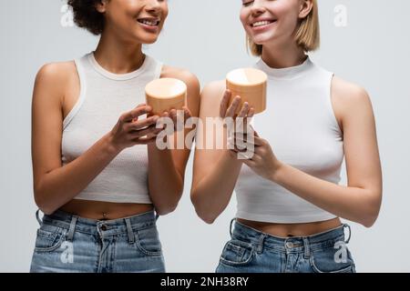 Zugeschnittene Ansicht von glücklichen Modellen aus verschiedenen Rassen in weißen Tanktops und Jeans mit Körpercreme isoliert auf grauem Stockbild Stockfoto