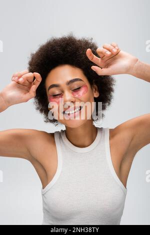 Fröhliche afroamerikanische Frau in weißem Tanktop und Kollagenaugen posieren mit geschlossenen Augen isoliert auf grauem Stockbild Stockfoto