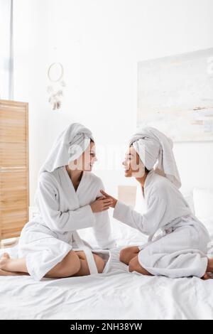 Fröhliche gemischtrassige Frauen in weißen Bademänteln und Handtüchern, die sich anschauen, während sie auf dem Bett sitzen, Stockbild Stockfoto
