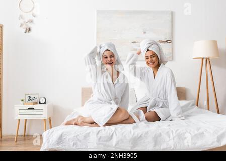 Fröhliche, multiethnische Frauen in weißen Bademänteln und Handtüchern, die auf dem Bett auf die Kamera starrten, Stockbild Stockfoto