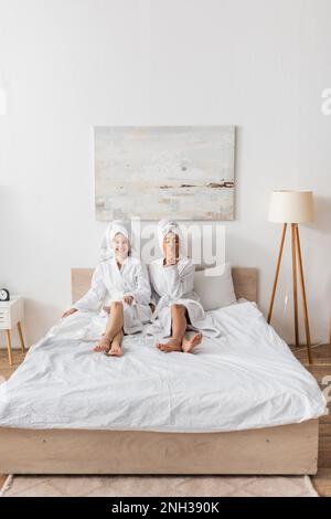 Volle Länge einer afroamerikanischen Frau, die in der Nähe eines fröhlichen Freundes einen Luftkuss bläst, im Bademantel und Handtuch auf dem Bett zu Hause, Stockbild Stockfoto