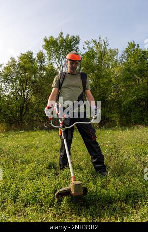 Junger erwachsener Mann in Schutzkleidung, der im Garten grünes Gras mit einem elektrischen Rasentrimmer mäht. Seitenansicht. Stockfoto