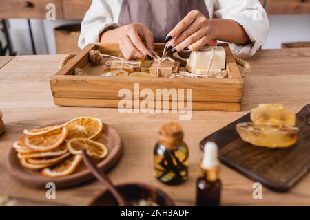 Kurzer Blick auf eine afroamerikanische Handwerkerin, die Garn auf handgefertigter Seife neben getrockneten Orangenscheiben und ätherischen Ölen auf verschwommenem Vordergrund, Stock Imag bindet Stockfoto