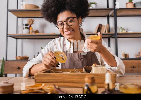Fröhliche afroamerikanische Handwerkerin in Brillen, die handgemachte Seife in der Nähe natürlicher Zutaten auf verschwommenem Vordergrund hält, Stockbild Stockfoto