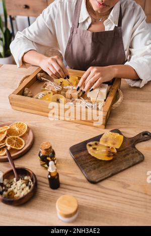 Freigestellte Ansicht einer afroamerikanischen Frau, die Garn an der Seifenleiste in der Nähe von ätherischen Ölen und getrockneten orangefarbenen Scheiben in einer Kunsthandwerkstatt bindet, Stockbild Stockfoto