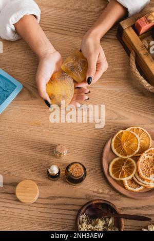 Draufsicht auf gekürzte afroamerikanische Handwerkerin mit hausgemachter Seife neben getrockneten Orangenscheiben und ätherischen Ölen, Stockbild Stockfoto