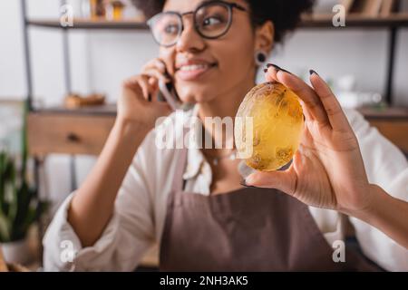 Handgefertigte Seife in der Hand einer frohen afroamerikanischen Handwerkerin, die die Bestellung auf dem Smartphone auf verschwommenem Hintergrund annimmt, Stockbild Stockfoto