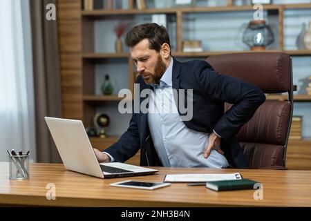 Ein erwachsener Mann, der in einem modernen Büro mit einem Laptop arbeitet, ein leitender Chef mit starken Rückenschmerzen, ein Geschäftsmann am Arbeitsplatz, der überlastet arbeitet und sich den Rücken mit der Hand massiert, starke Schmerzen beim Sitzen. Stockfoto