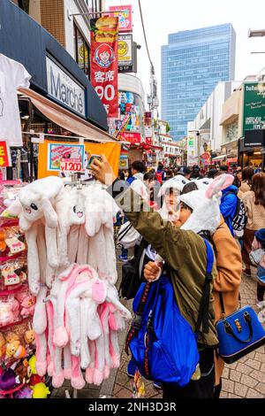 Chinesische Touristen machen ein Selfie, während sie weiße Kaninchenmützen tragen, die von Ausstellungsanzeige voll von ihnen draußen Geschäft mit Hintergrund von Takeshita Dori, Harajuku stehen. Stockfoto