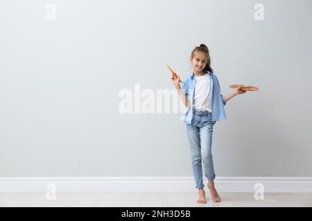 Kleines Mädchen mit Pinsel und Farbpalette in der Nähe von hellen Wänden in Innenräumen. Platz für Text Stockfoto