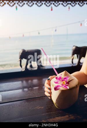 Frau trinkt süße frische Kokosnuss mit Stroh im Restaurant mit hölzernen Elefanten Dekoration am Strand mit Blick auf das Meer bei Sonnenaufgang. Stockfoto