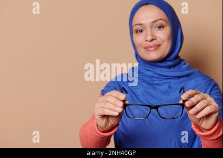 Selektiver Fokus auf schwarze Brillen - Brille in den Händen stilvoller muslimischer Frauen, isoliert auf beigem Hintergrund Stockfoto