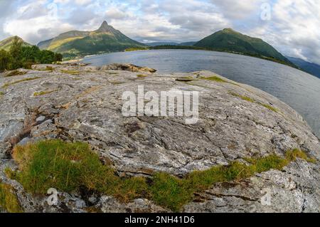 Eine zerklüftete felsige Küste mit grünem Gras überblickt das ruhige Wasser, umgeben von majestätischen Bergen unter einem bewölkten Himmel in Norwegen. Stockfoto