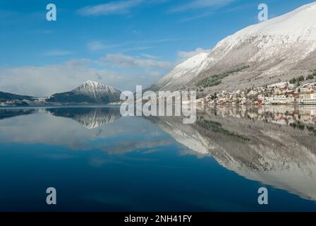 Schneebedeckte Berge und ein friedliches Dorf spiegeln sich perfekt im stillen Wasser an einem klaren Wintertag wider. Stockfoto