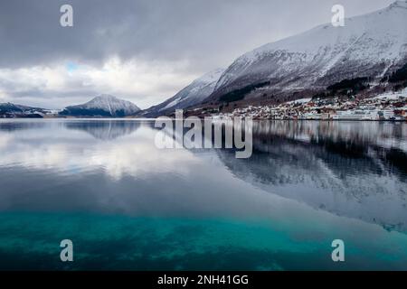 Eine ruhige Winterszene bietet schneebedeckte Berge, die sich im ruhigen Wasser neben einem malerischen Dorf in Norwegen spiegeln. Stockfoto