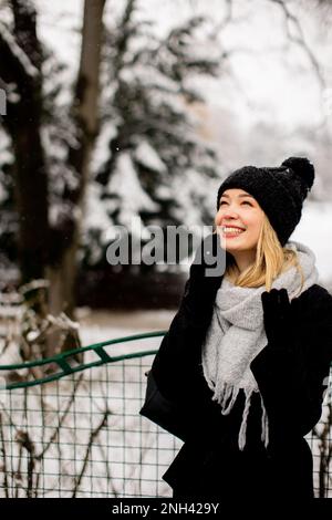 Eine junge Frau, die warme Winterkleidung trägt und eine Strickmütze trägt, lächelt glücklich, wenn sie im Schnee steht und ihr Handy benutzt Stockfoto