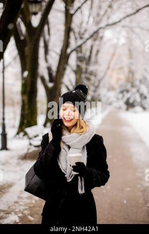 Eine junge Frau, die warme Winterkleidung trägt und eine Strickmütze trägt, lächelt fröhlich, wenn sie im Schnee steht und mit ihrem Handy einen Kaffeebecher hält Stockfoto