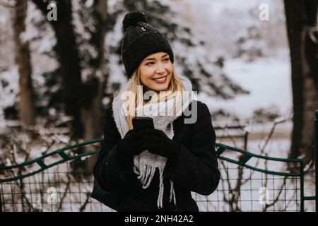 Eine junge Frau, die warme Winterkleidung trägt und eine Strickmütze trägt, lächelt glücklich, wenn sie im Schnee steht und ihr Handy benutzt Stockfoto