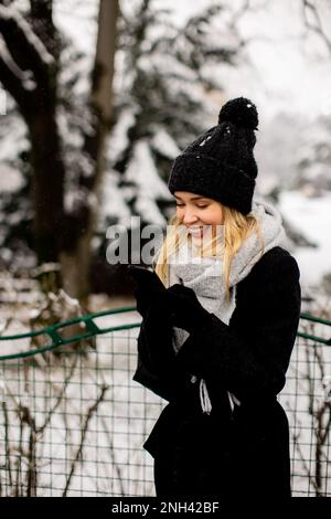 Eine junge Frau, die warme Winterkleidung trägt und eine Strickmütze trägt, lächelt glücklich, wenn sie im Schnee steht und ihr Handy benutzt Stockfoto