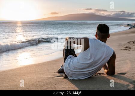 Ein Mann auf dem Rücken, der auf dem Sand liegt und den Sonnenuntergang am Strand beobachtet. Reisen Stockfoto