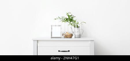 Leerer Bilderrahmen, Nest mit Ostereiern, Häschen, grüne Äste auf der Kommode nahe der weißen Wand. Banner für Design Stockfoto