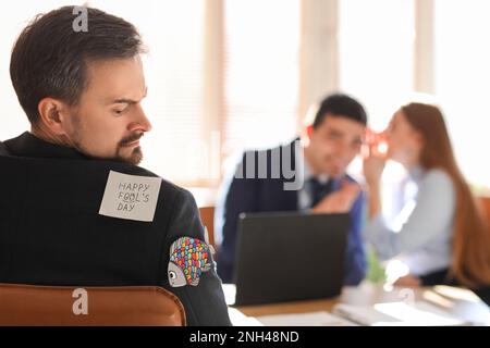 Haftnotiz und Papierfisch auf dem Rücken eines wütenden jungen Mannes im Amt. Feier zum 1. April Stockfoto