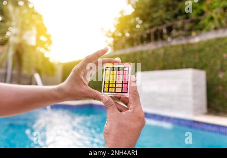 Hand hält einen pH-Wert- und Chlortester, Hand zeigt Wassertestkit mit Hintergrund des Schwimmbeckens. Wartungspersonal mit einem Pool-ph und Stockfoto