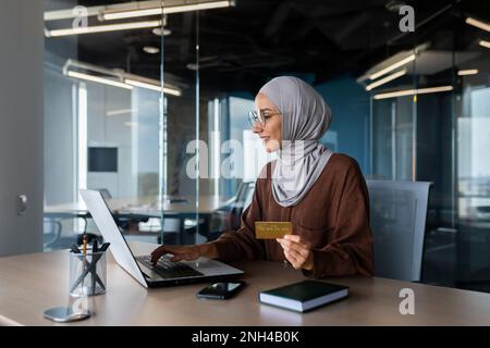 Erfolgreiche muslimische Frau im Hijab bei der Arbeit in modernen Büros, Geschäftsfrau mit Bankkreditkarte und Laptop kauft online im Online-Shop ein, zufriedene und lächelnde Frau am Arbeitsplatz. Stockfoto