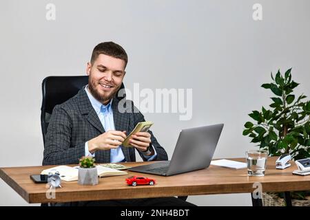 Ein junger, bärtiger Geschäftsmann, der an einem Laptop arbeitet und Geld hält Stockfoto