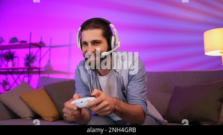 Ein fröhlicher junger Mann mit Headset spielt Videospiele auf der Konsole, während er auf der Couch vor dem Fernseher sitzt. Gamer, die zu Hause Spaß haben Stockfoto