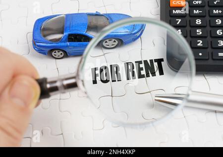 Ein Mann schaut durch eine Lupe auf weiße Puzzles mit der Inschrift - ZUR MIETE. Auf den Rätseln befinden sich ein blaues Automodell, ein Taschenrechner und ein Stockfoto