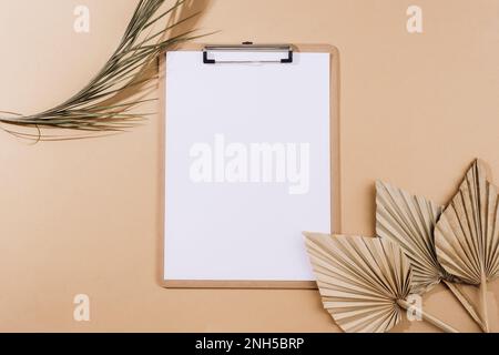 Schreibtisch mit leerem Klemmbrett, Fächerblättern und getrockneten Palmenblättern auf beigefarbenem Hintergrund. Draufsicht, flaches Lay, Modell. Stockfoto