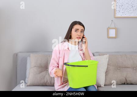 Junge Frau ruft Dachreparaturservice an, während sie zu Hause das Wasser aus der Decke sammelt Stockfoto