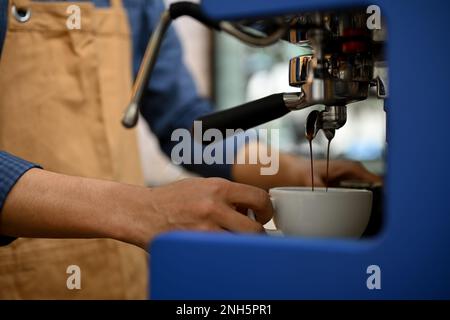 Ein männlicher Barista in der Schürze bereitet Kaffee für seine Gäste zu und macht Espresso Shot aus der Espressomaschine. Seitenansicht, Nahaufnahme Stockfoto