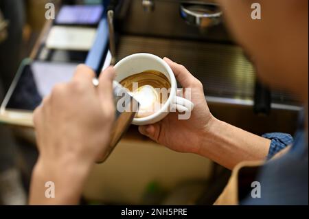 Nahaufnahme eines talentierten männlichen asiatischen Baristas, der aufgeschäumte Milch in eine Tasse gießt, Latte Art herstellt und im Café arbeitet. Stockfoto