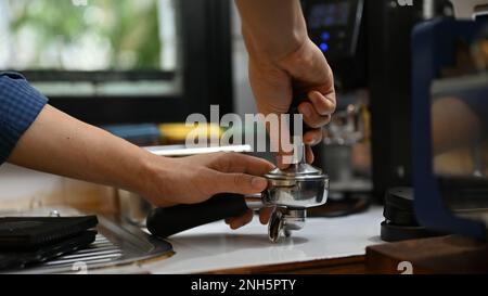 Ein professioneller männlicher Barista hält den Portafiler, presst Kaffeepulver mit einem Kaffeetampfer und bereitet die Bestellung des Gastes im Café vor. Nahaufnahme Stockfoto