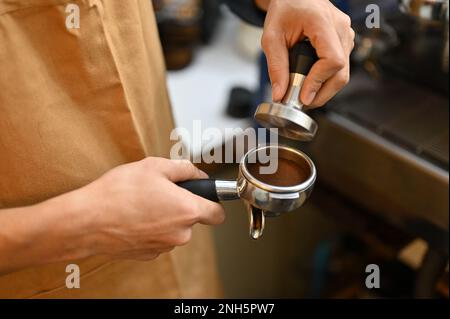 Ein professioneller männlicher Barista hält den Portafiler, presst Kaffeepulver mit einem Kaffeetampfer und bereitet die Bestellung des Gastes im Café vor. Café oder Café Stockfoto