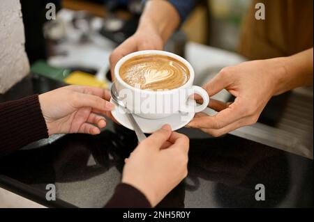 Nahaufnahme eines männlichen Baristas, der einem Gast im Café eine Tasse Latte überreicht. Stockfoto