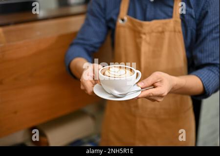 Ein männlicher Barista oder Kellner hält eine Tasse wunderschöner Latte Art vor der Theke im Café. Zugeschnittenes Bild Stockfoto