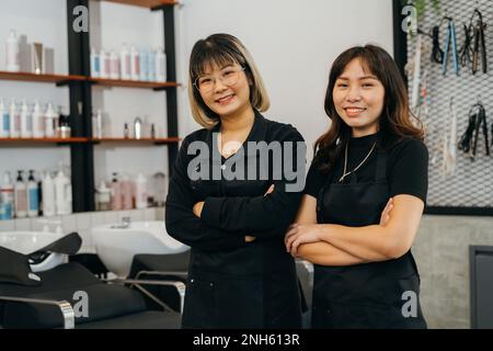 Porträt eines Teams glücklicher, erfolgreicher junger asiatischer Friseurin, die mit Schürze und Brille mit gefalteten Händen lächelnd in der Kamera steht Stockfoto